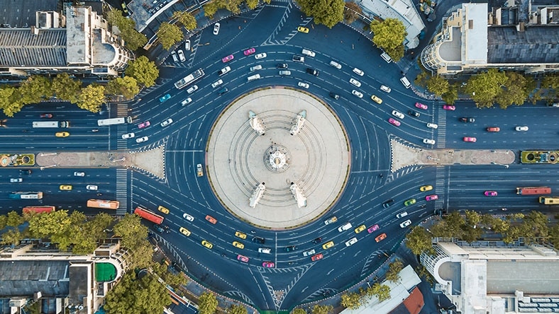 Aerial view of a large roundabout with multiple roads, vehicles, surrounding buildings, and greenery.