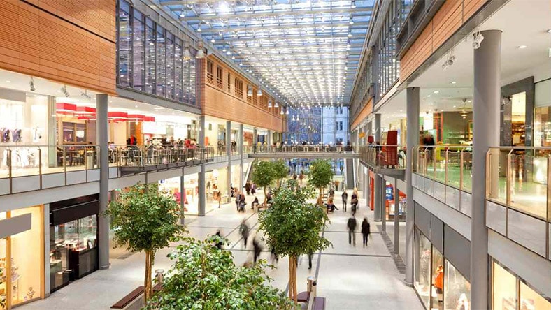 Modern multi-level shopping mall interior with storefronts, indoor trees, and people walking through a bright atrium.