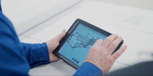 Una imagen en primer plano de manos sosteniendo una tableta. La tableta muestra una renderización de varios edificios. 