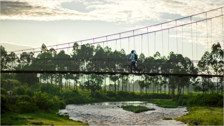 A side-angle view of a Bridges to Prosperity trail bridge on a mostly sunny day. A person on a bicycle is crossing the bridge, roughly halfway across; the bridge stretches left to right above a flowing river, with lush greenery on both river banks. Background: A line of tall trees with large green canopies in the far distance.