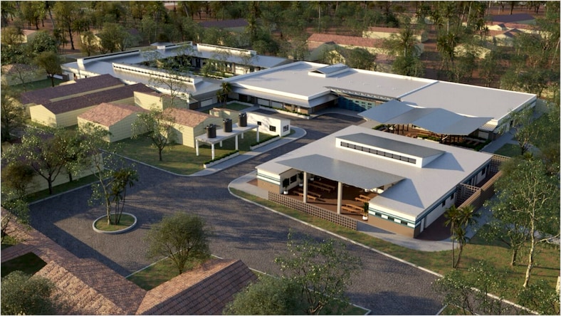 Rendering of an aerial view of Build Health International's Maternal Center of Excellence in Sierra Leone. A paved driveway and parking lot leading to a large 1- to 2-story building complex with a light gray roof on a sunny day. The buildings are surrounded by various trees and green grass.