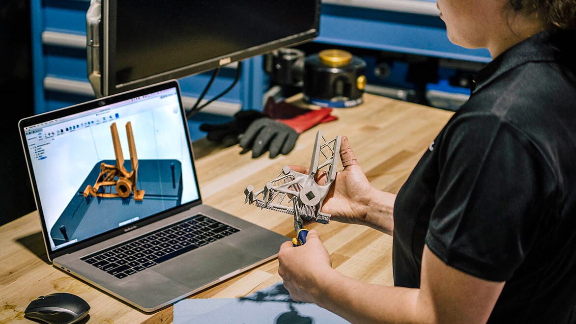 A woman holds a 3D printed prototype while sitting in front of a laptop with generative design software opened.