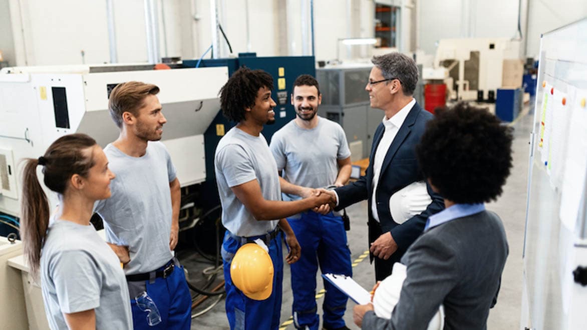 A group of 4 people watch as two men shake hands. Three of the people are holding hard hats.