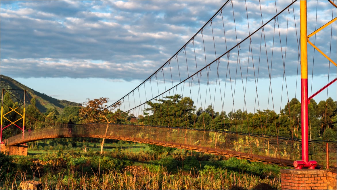 A brightly lit, landscape, side-angle view of a Bridges to Prosperity trail bridge on a partly cloudy day. A person on a bicycle is crossing the bridge. Background: A mountain extends upward to the left in the far distance; the area is surrounded by tall trees with large green canopies. Foreground: tall grasses, partly covered by shadows from surrounding trees.