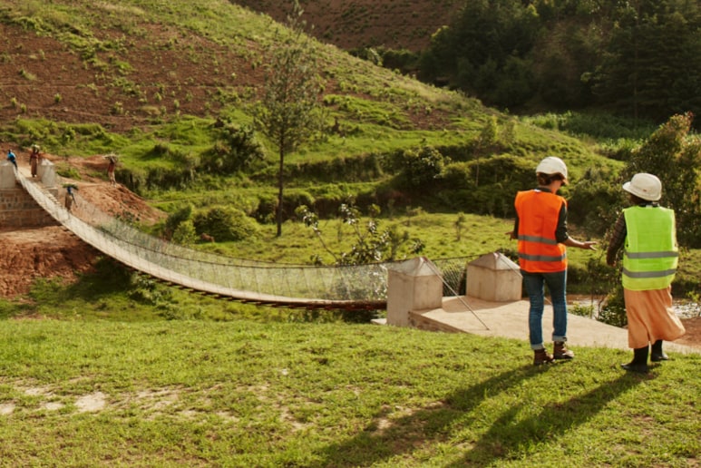 Two women wearing white hard hats and high-viz vests standing and talking next to a Bridges to Prosperity bridge along a grassy hillside on a sunny day.