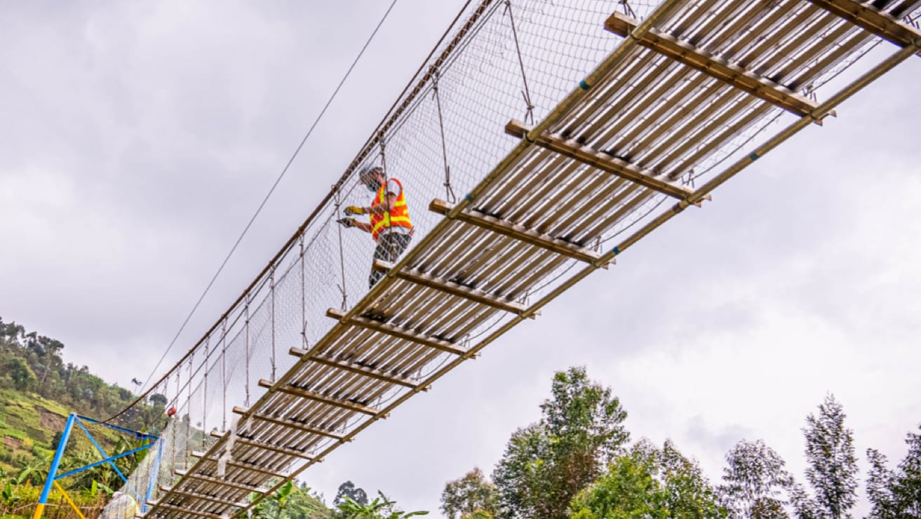 Ground view of a construction worker standing on a B2P suspension bridge against a backdrop of cloudy skies and green, forested hillside.