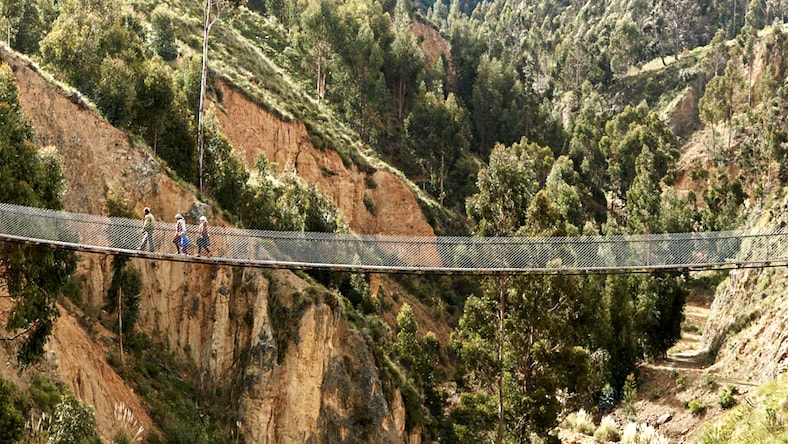 Aerial side-view of a Bridges to Prosperity trail bridge spanning left to right between two mountain sides on a sunny day. Three individuals are walking along the bridge, toward the left side of the bridge in the image. Surrounding terrain is rocky and steep with tall trees growing through the rocks into the distance.