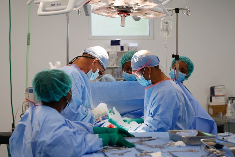 Operating room with two surgeons and three technicians wearing scrubs, masks, and hair coverings.
