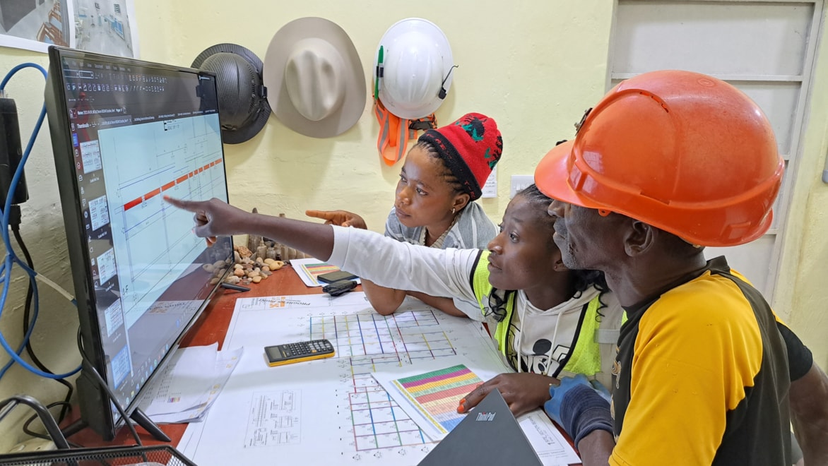 Left-side view of three individuals—two women and one man—sitting a desk looking at a document on a large computer monitor together. The man, closest to the camera, is wearing an orange hardhat and bright yellow shirt. The woman in the middle is wearing a high-viz yellow vest, pointing at an item on the screen, with eyebrows raised; the woman to her right is wearing a red beanie, resting her elbows on the desk.