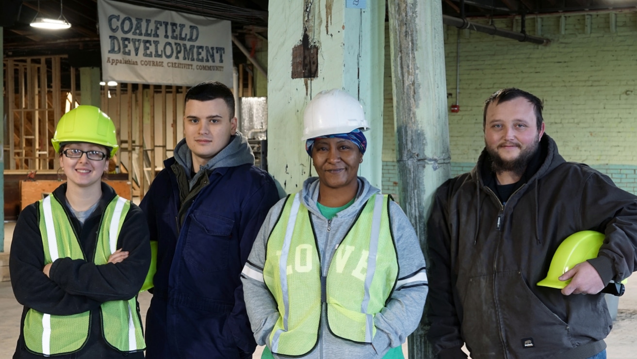 Four Coalfield Development trainees smiling and posing for a group photo — two women wearing hard hats and hi-viz vests, and two men wearing hoodies.