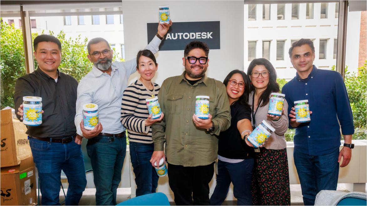 Group of Autodesk employees standing indoors, smiling and holding jars in front of an Autodesk sign.