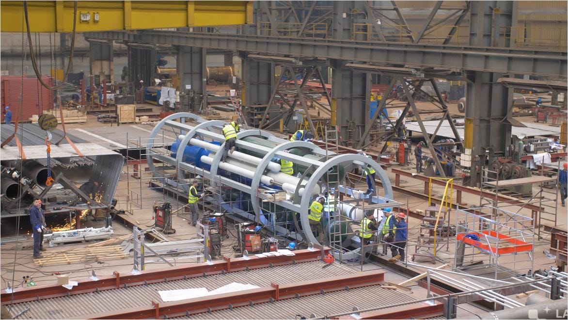 Construction workers at Last Energy assembling a modular nuclear reactor—large cylindrical machinery—inside an industrial facility, surrounded by scaffolding, tools, and equipment.