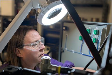 A Found Energy team member wearing safety glasses works under a magnifying lamp, inspecting electrical components inside a prototype system.