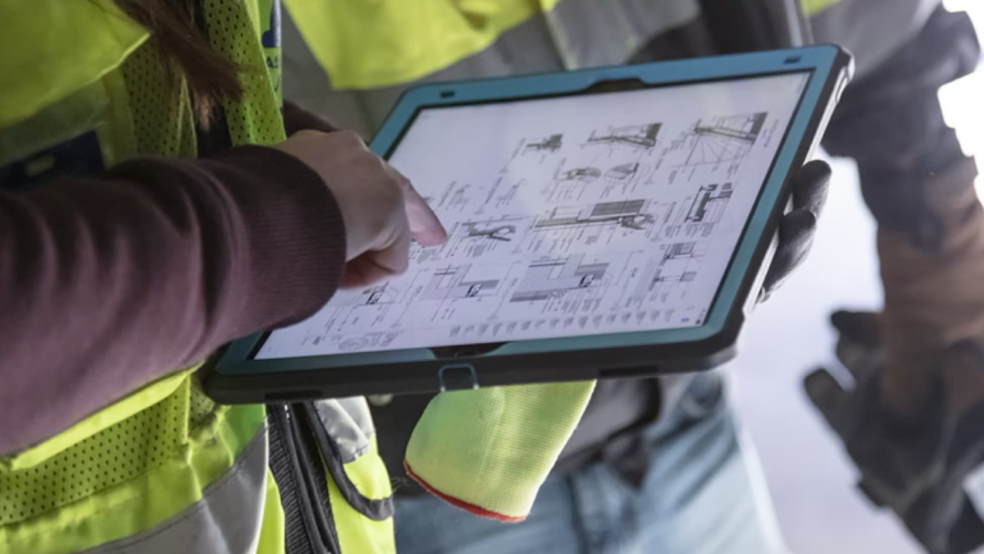 Close-up view of construction worker wearing high-viz yellow vest pointing at schematics on iPad screen. A fellow construction worker stands to main subject's left, also wearing high-viz yellow vest and work gloves.