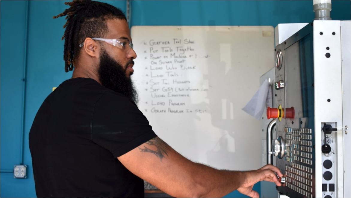 Side view of Adonis Summerville operating a CNC mill at JARC Chicago. Summerville is wearing safety glasses and a black t-shirt, reaching out with his right hand to touch the controls of a CNC milling machine. A whiteboard hangs on a light-blue wall behind him.
