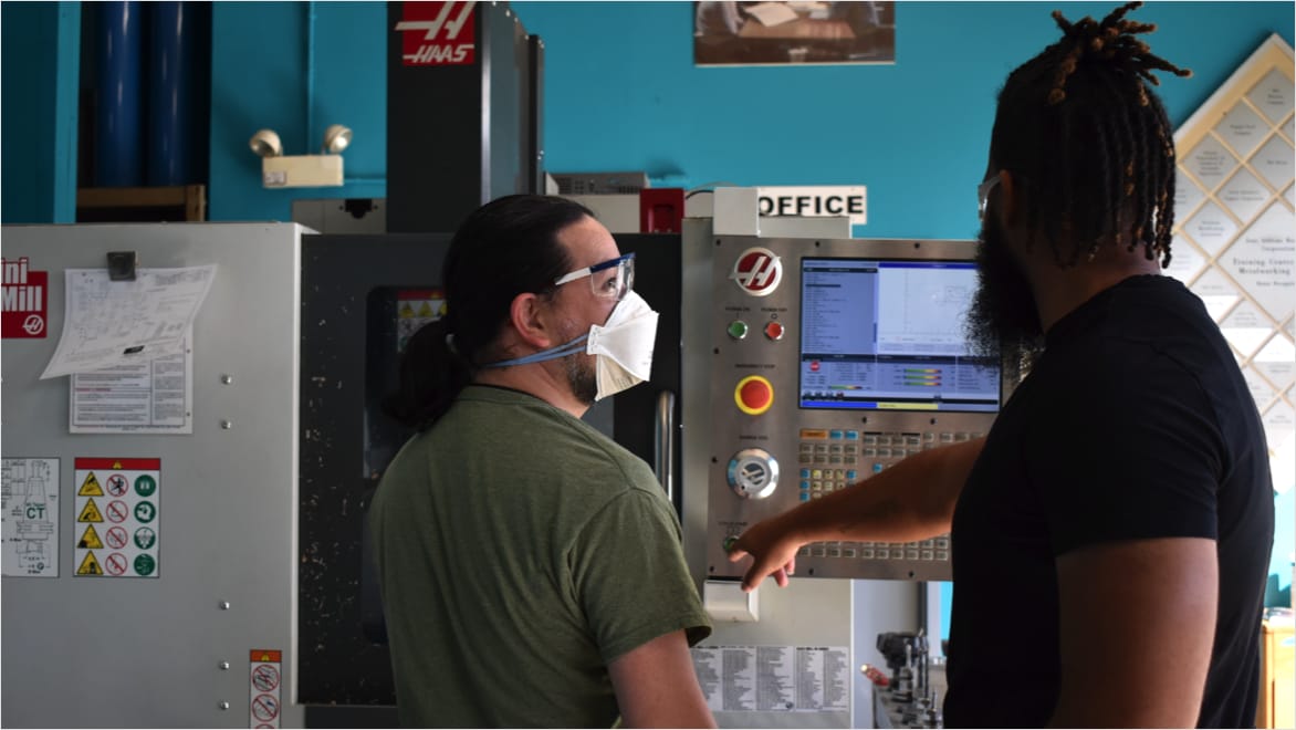 Over-the-shoulder view of Adonis Summerville (right) teaching a JARC trainee (left) how to operate a Haas Mini Mill CNC milling machine. Summerville extends his right hand to touch a button on the control panel as the trainee, wearing safety glasses and a face covering, looks up at him. The CNC machine takes up the majority of the background.
