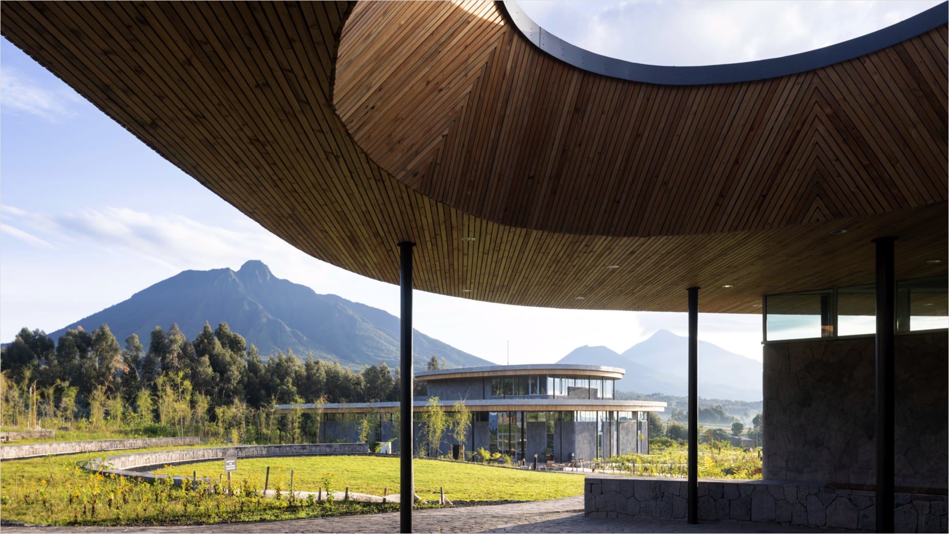 Ground view from the patio of one of the buildings of The Ellen DeGeneres Campus of the Dian Fossey Gorilla Fund in Rwanda. Foreground: Stone patio ground with curved, wood-paneled roof with large, round opening in the top, supported by thin black poles. Background: Another campus building in the distance, surrounded by green grass, a stone retaining wall, and a thick row of trees, with a large mountainous range in the background — sunny skies with scattered clouds.