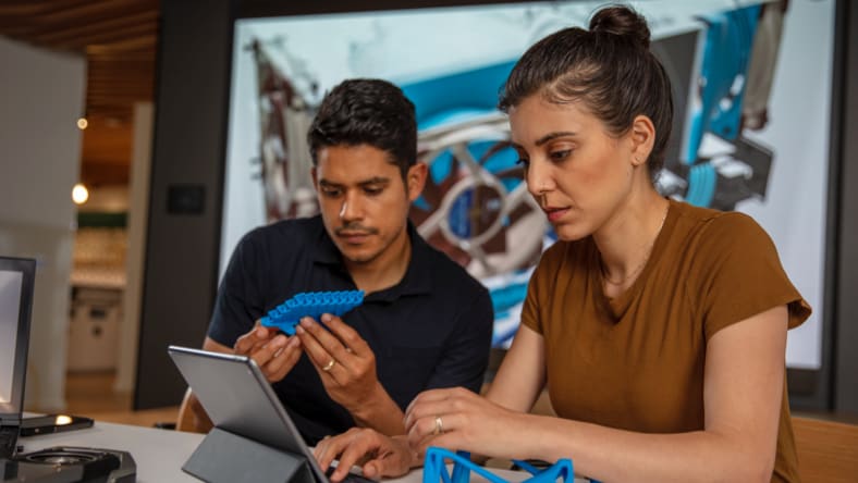 A man and a woman sitting at a table looking at blue 3D-printed objects while consulting Autodesk software on a tablet.