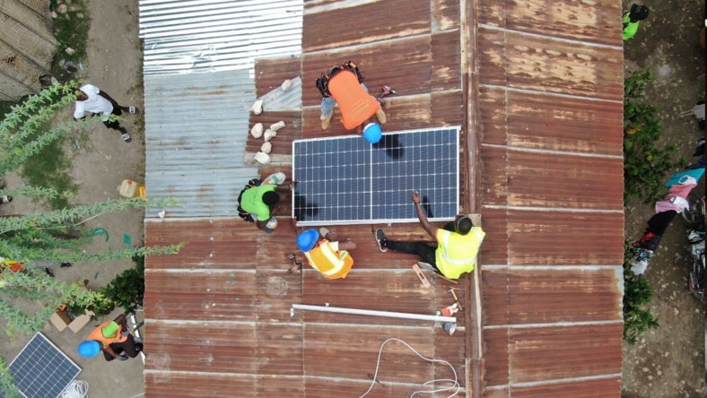 Arial view of four workers installing solar panels on rust-colored metal roof.