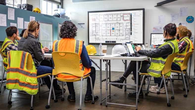 A group of construction workers wearing high-viz yellow vests sit at a horseshoe-shaped table arrangement in a brightly lit room, looking at a flatscreen TV displaying a black-and-white floor plan.