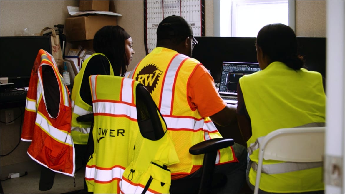 Three individuals wearing high-viz vests sitting together at a desk, looking at computer being operated by Marcus Lee (center), as he demonstrates AutoCAD fundamentals to the two Revolution Workshop trainees on either side of him.