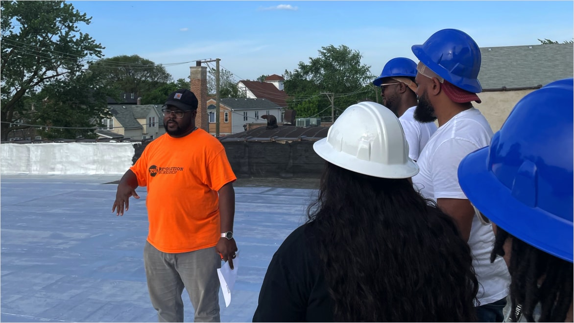 Marcus Lee (left), wearing a bright orange Revolution Workshop t-shirt and a black baseball cap, explains the process of getting a roof estimate to four Revolution Workshop trainees who are all wearing t-shirts and hard hats. The group is standing on a roof on a sunny day, with trees and other buildings in the background.