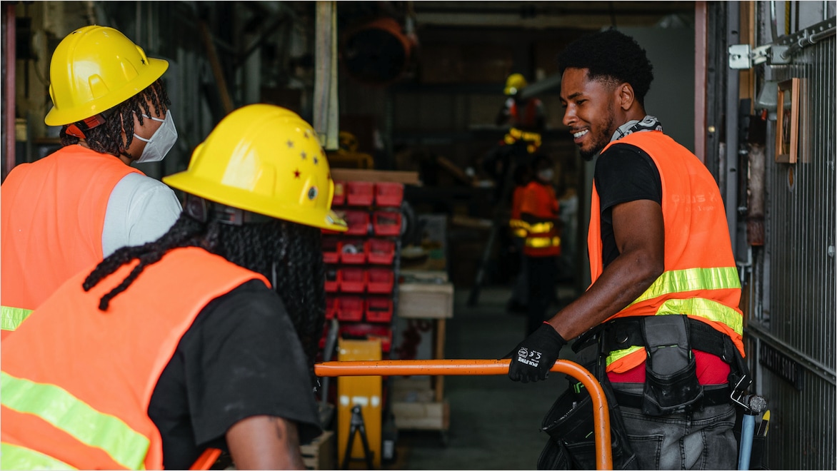 Three young men—two wearing yellow hard hats facing away from the camera, one facing toward the camera, smiling and wearing work gloves—tag-team moving an orange cart through a warehouse. All are wearing orange and yellow hi-viz safety vests.