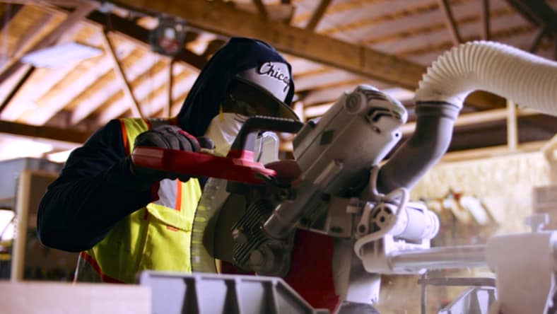 A Revolution Workshop trainee wearing a high-viz yellow vest, black sweatshirt, a "Chicago" baseball hat, and face covering operating a miter saw at Revolution Workshop in Chicago. Background: tall, vaulted ceiling with visible rafters and sunlight shining in.