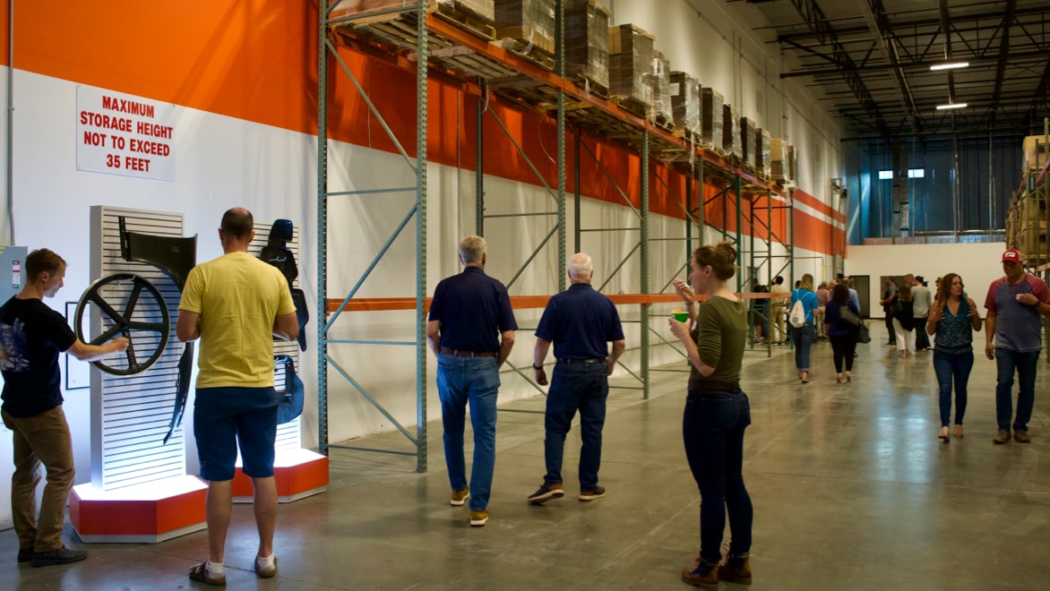 Attendees of the Pecos facility ribbon-cutting touring the new space. Roughly a dozen individuals of various ages and demographics walking along the warehouse wall. Two men looking at the carbon fiber bicycle rim on display, toward the left of the photo.