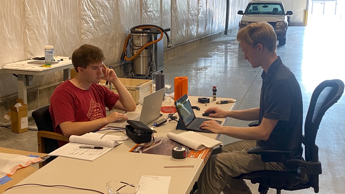 Two Vartega interns working at computers on a folding table at the new Pecos facility in Denver, Colorado. Both individuals are wearing t-shirts and looking at their computers. The table is covered with writing utensils, notebooks, two reusable water bottles, and power cables. 