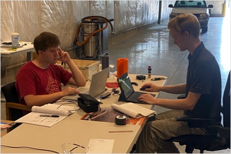 Two Vartega interns working at computers on a folding table at the new Pecos facility in Denver, Colorado. Both individuals are wearing t-shirts and looking at their computers. The table is covered with writing utensils, notebooks, two reusable water bottles, and power cables. 