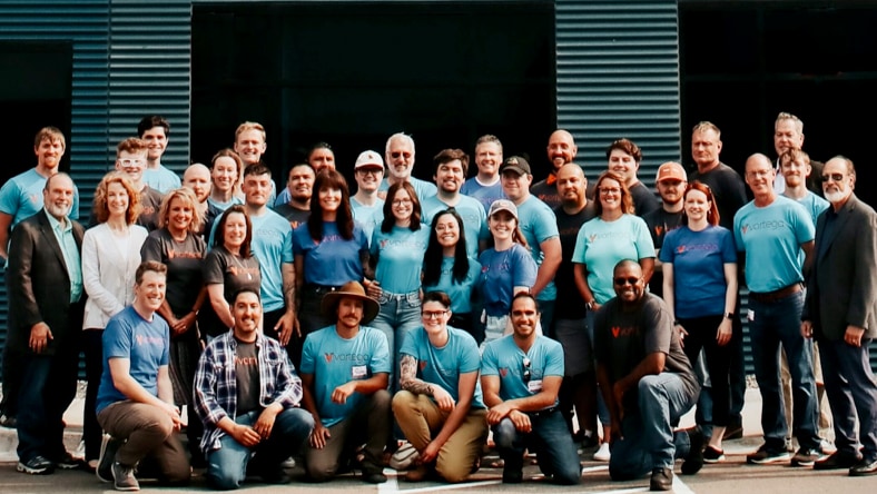 Group photo of the Vartega team and partners—roughly 40 individuals, many wearing light-blue Vartega-branded t-shirts, smiling and posing for the shot in front of Vartega's new Pecos facility in Denver, Colorado.