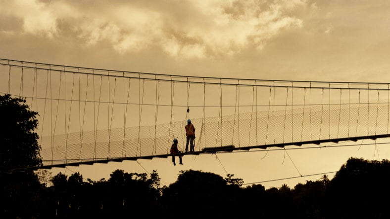 Side view, from a distance, of a silhouetted B2P suspension trail bridge with two construction workers in the middle of it (one sitting, one standing). Trees line the base of the frame, under the bridge and workers, with a backdrop of a partly cloudy sky at dusk.