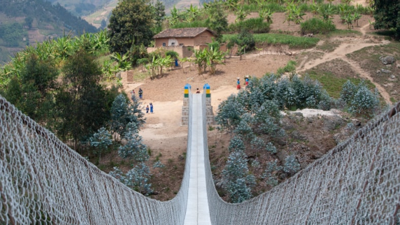 Aerial view from one end of a long B2P trail bridge, with chain-link fencing on either side of the bridge, roughly four feet heigh. In the distance, at the other end of the bridge: a house sitting atop a grassy hill surrounded by lush trees and shrubs, with more mountains and trees in the distance, around and behind the 2-story hilltop house.