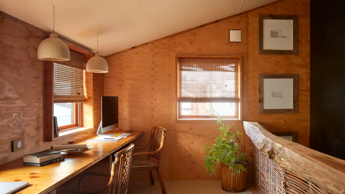 An eye-level view of the interior of the BLDUS Grass House project, with MushLume pendant lamps on the left side of the room, and BamCore bamboo panel walls on the far and left-side walls. An iMac, a laptop, a short stack of books, and a notepad with a pen atop a long wooden desk along the left side of the photo; three empty wooden chairs are placed alongside the desk.