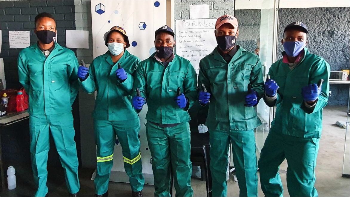 A group of five BluLever Education apprentices posing for a photo, wearing matching grey canvas uniforms, face coverings, and blue work gloves. The four individuals on the right are giving double thumbs-ups and wearing hats (one bucket, three baseball). The group is standing in BluLever's training facility, with brick and concrete walls in the background, and a large white paper easel with the words "Our Norms" visible.