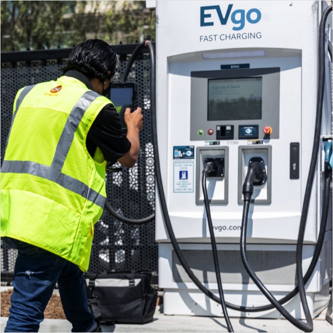 A man with slicked-back black hair, wearing a black face covering, hi-viz yellow vest, black t-shirt, and blue jeans—standing with his back to the camera in front of an EVgo-branded EV charging station on a sunny day. The man is using both hands to hold up a tablet-sized device, facing the EV charging station as though taking a picture of it.