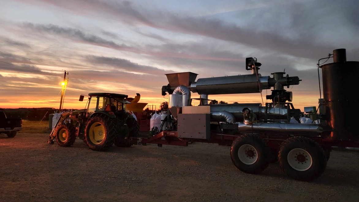 Side-view of tractor towing Applied Carbon's biochar technology unit on a trailer over flat dirt ground in an open field at dusk, with a colorful sunset in the background.