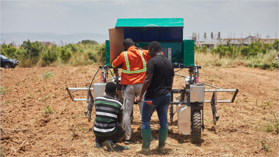 A candid shot of a person riding a motorcycle on a dirt road sunny day. The man is wearing a black motorcycle helmet and has a beige bag hanging across his chest, on his right side. The background is blurred, with palm fronds hanging above and behind the motorcyclist.