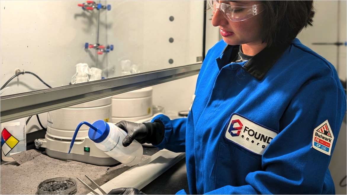 Found Energy Senior Materials Engineer Ingrid Guha standing in front of a venting workbench, spraying water from a translucent squirt bottle onto an aluminum feedstock (in a petri dish) that has been activated with Found Energy's catalyst to study its reactivity. Guha is wearing a blue Found Energy branded blue collared shirt, clear safety classes, and black nitrile gloves.