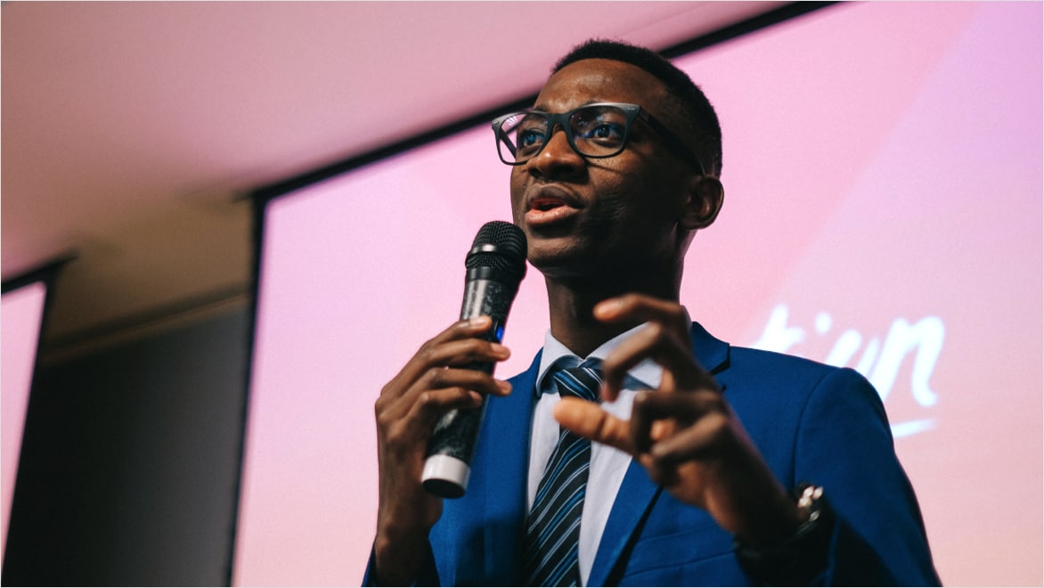 Close-up candid/action shot of a young man wearing a blue blazer, glasses, button-down shirt and a striped blue tie, holding a microphone in his right hand and his left hand in front of him, fingers curled as if grabbing something, presenting at a Generation workshop or event.