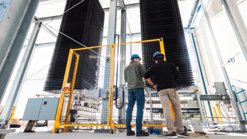 Two Heirloom engineers wearing hardhats, facing away from the camera, standing in front of Heirloom's direct air capture unit — two tall black square columns surrounded by metal scaffolding and caging around the base of the technology. Bright, sunny day.