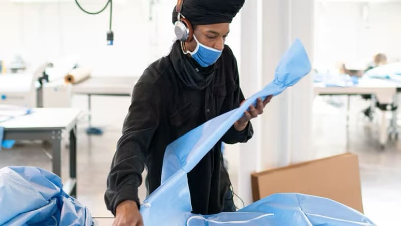 A person wearing a black button-down shirt, headphones, and a blue face covering, holding and examining a wide strip of blue PPE material in a textile workshop.