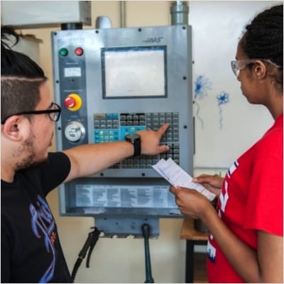 Over-the-shoulder photo of two JARC Rhode Island trainees standing in front of a machine. Individual on the left pushes a button on a keypad while the individual on the right looks on and takes notes.