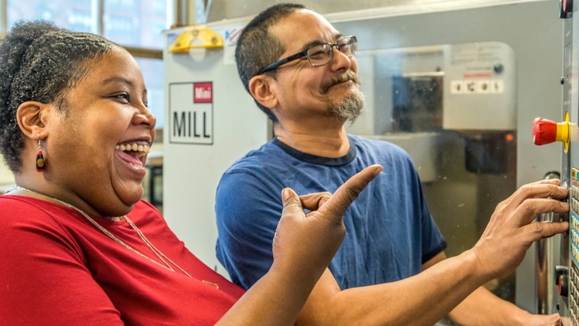 A woman and a man stand in front of a CNC machine, both smiling. The man has both hands on the machine, pressing buttons as they watch it work.