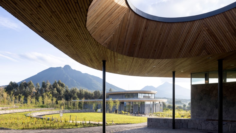 Ground view from the patio of one of the buildings of The Ellen DeGeneres Campus of the Dian Fossey Gorilla Fund in Rwanda. Foreground: Stone patio ground with curved, wood-paneled roof with large, round opening in the top, supported by thin black poles. Background: Another campus building in the distance, surrounded by green grass, a stone retaining wall, and a thick row of trees, with a large mountainous range in the background — sunny skies with scattered clouds.