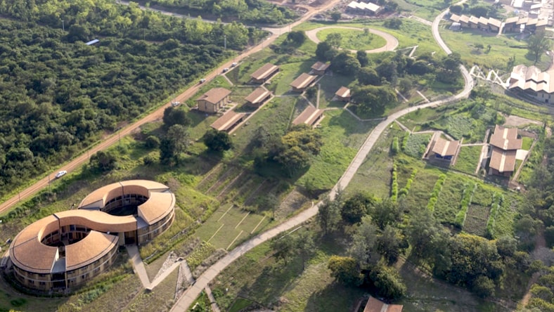 Aerial drone photo of the MASS RICA campus, with the main infinity-shaped building in the lower-left corner, with a series of buildings appearing intermittently on the campus grounds. The campus is surrounded by forest and includes what appear to be crop fields. A slightly windy road weaves through the campus, connecting the main building to the others.