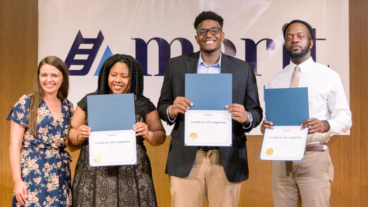 Four individuals—two women and two men—holding Merit America certificates of completion in front of a large Merit America sign.