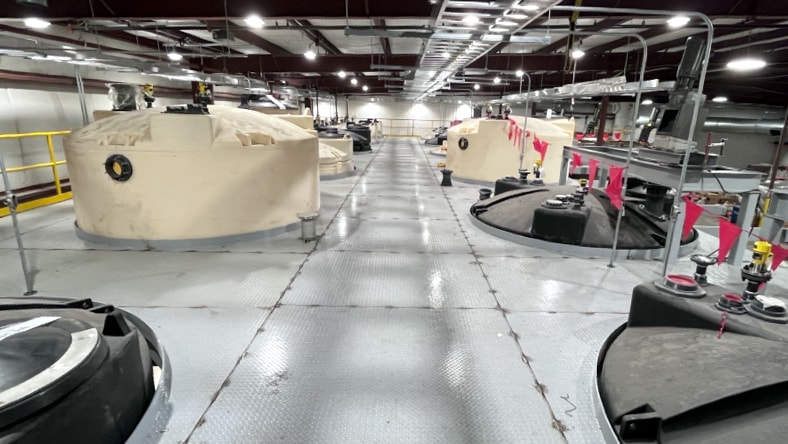 A top view of Nth Cycle’s Oyster system — a long array of large metal, cylindrical holding tanks in an expansive, well-lit warehouse space with paneled, sheet-metal flooring.