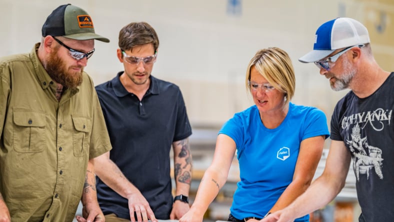 Pallet CEO and Founder Amy King, wearing a blue Pallet-logoed t-shirt, pointing at a schematic on a table, with two Pallet employees two her right and one to her left (three men). All four individuals are wearing safety glasses and looking intently at the schematic design.